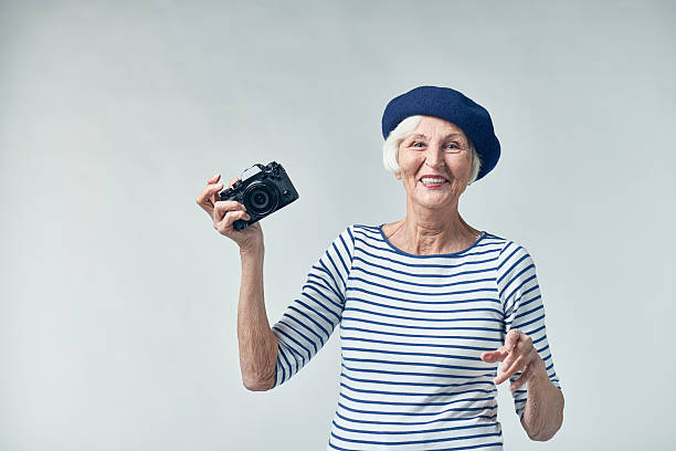 Waist-up portrait of cheerful senior woman in stylish clothes holding camera in hand and standing against white background