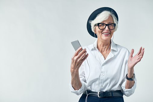 Joyful elderly woman in hipster clothes listening to music on smartphone and looking away with wide smile