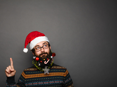 handsome man with decorated christmas beard in red santa claus hat gesturing upwards with his finger to copy space above his head over grey background