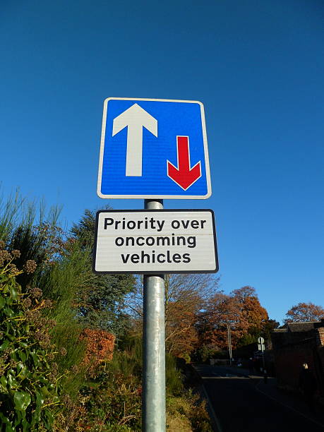 'Priority over oncoming vehicles' road sign 'Priority over oncoming vehicles' road sign. Rectangular Department for Transport information sign. Blue with white border, with white and red arrows indicating appropriate traffic priority. Roadside hedge and blue sky in background. uk road signs stock pictures, royalty-free photos & images