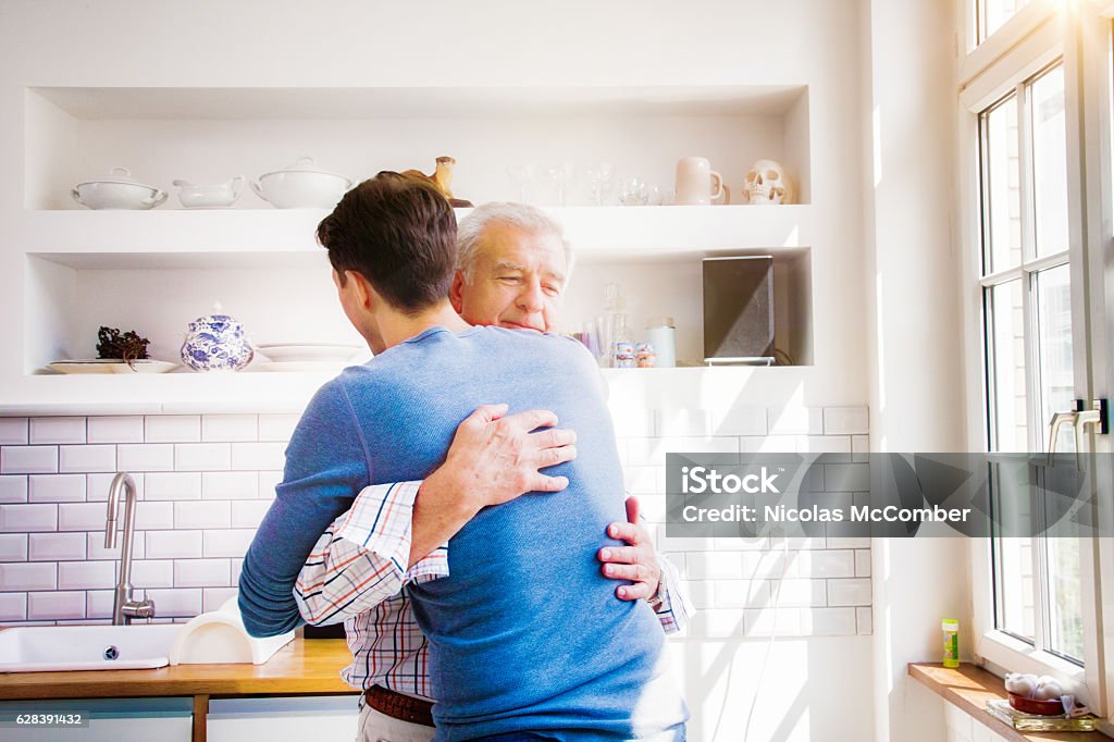シニアの男は日当たりの良いキッチンで成熟した息子を抱きしめる - 許しのロイヤリティフリーストックフォト