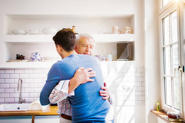 Senior man embraces mature son in sunny kitchen Senior man embraces mature son in sunny kitchen with a proud smile. mercy hug stock pictures, royalty-free photos & images