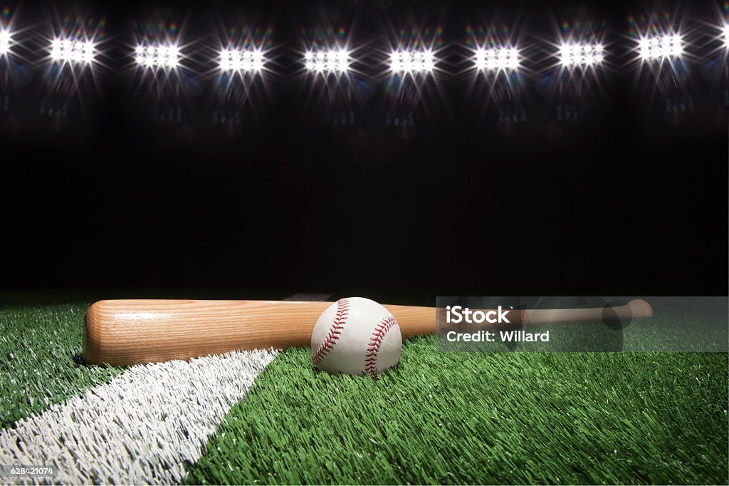 Baseball and bat at night under stadium lights Baseball and bat at night under stadium lights on grass field with white stripe Baseball - Sport Stock Photo