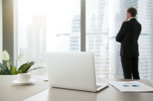 Company president in formal suit standing, looking through window at cityscape, dreaming, planning new projects, resting after intensive work. Focus on laptop on office table. Business success concept