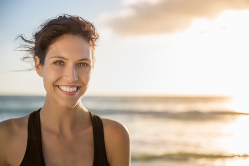 Portrait of happy female athlete at beach. Confident young woman is against sky during sunset. She is sportswear.