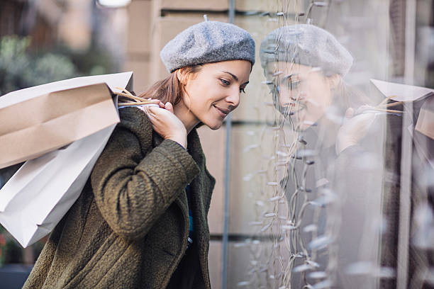 Smiling woman doing window shopping Side view of smiling woman looking into shop window. Mid adult female is wearing winter coat and flat cap while window shopping. She is carrying shopping bags. flat cap stock pictures, royalty-free photos & images