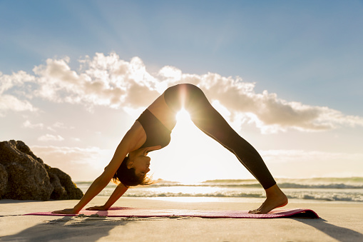 Side view of female athlete doing yoga in downward facing dog position on shore. Sunlight is streaming through young woman exercising at beach during sunset. She is in sportswear.