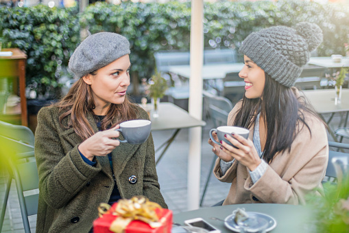 Happy female friends talking at sidewalk cafe. Women are looking at each other while holding coffee cups at table. Both are in warm clothing.