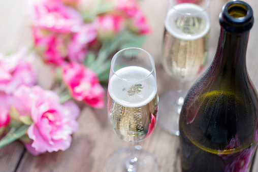 Bottle of Prosecco and two champagne glasses on a rustic garden picnic table with blossom or flowers in the background. Springtime or Summertime.