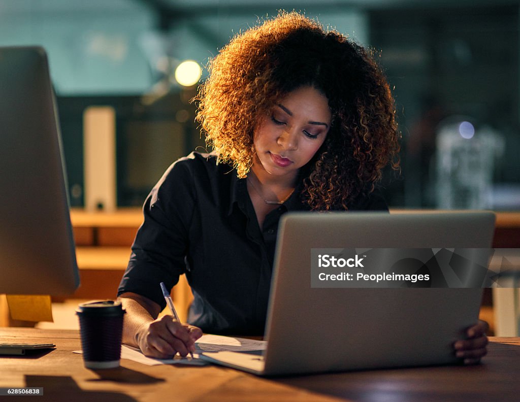 Being the best requires hard work and dedication Shot of a young businesswoman using a laptop and writing notes during a late night at work Working Stock Photo