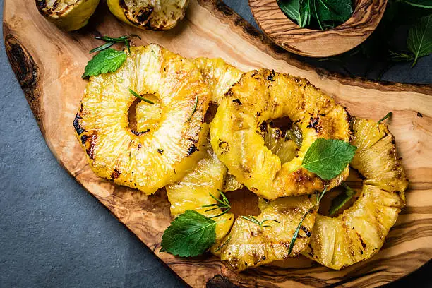 Grilled pineapple slices with fresh mint on olive wooden cutting board, gray slate background. top view