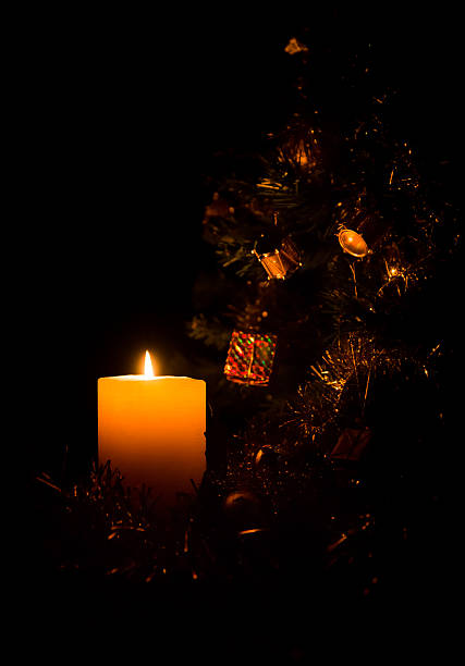 Glowing festive candle illuminating a Christmas tree and decorations. With a black background. Low light atmospheric night time scene.
