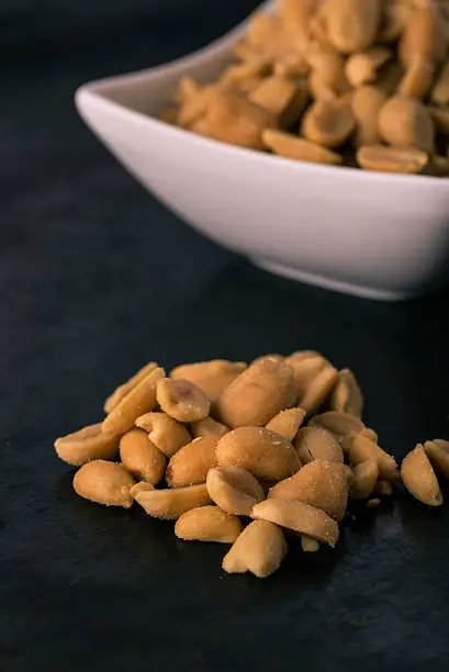 Vertical photo of salted peanuts. Light brown nuts with salt placed on heap in front of white square bowl full of others. Nuts placed on dark black baking tray board with worn surface.