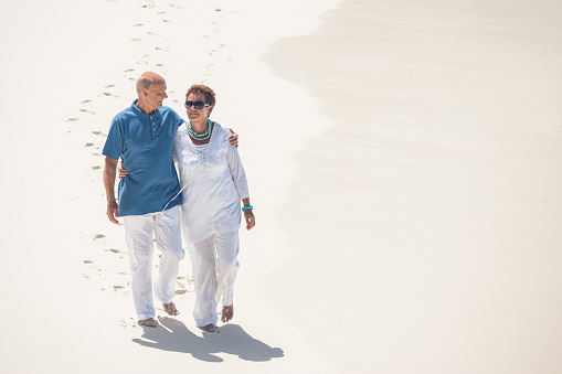 casual dressed best ager couple hugging walking barefoot on white sandy beach