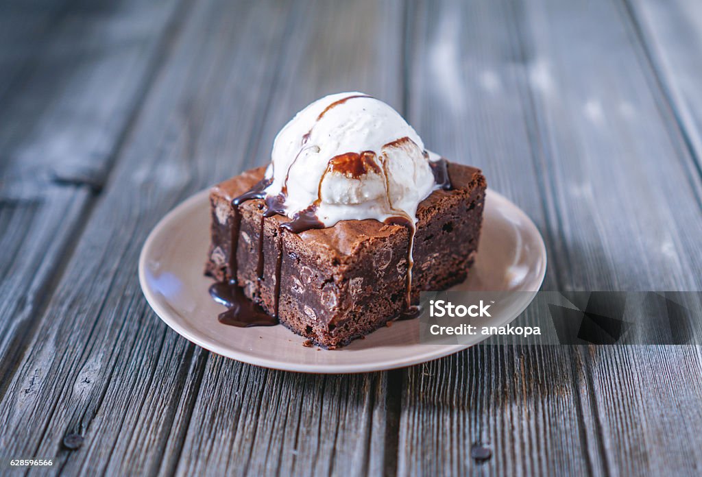 Chocolate Fudgy Brownie with Vanilla Ice Cream on top. Chocolate Fudgy Brownie with Vanilla Ice Cream and Chocolate Syrup on Wood Background. Selective focus, toning. Brownie Stock Photo