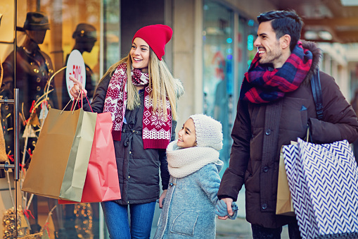 Young, happy couple and their daughter are looking in the shop window at Christmas