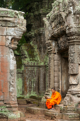 Cambodian monk in an orange robe sits on the ancient steps of a temple ruin and reads a book in a quiet moment, Siem Reap, Cambodia, Asia