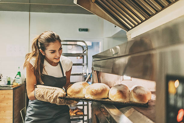 Baker pulling a tray with hot bread Young woman baker taking out the hot bread from the oven. artisanal food and drink stock pictures, royalty-free photos & images