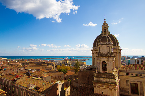 Catania, Sicily: Beautiful old town panorama with vibrant orange tile roofs; the cupola and clock tower of Duomo di Sant'Agata is in the foreground and a turquoise sea (and Catania's harbor) and blue sky (with a puffy cloud) in the background. Copy space in the sky. The baroque Cathedral of Saint Agatha was constructed in the 18th century.