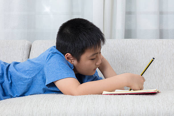 gelangweilte asiatische chinesische kleine junge schreiben buch auf dem sofa - reading and writing little boys reading asian ethnicity stock-fotos und bilder