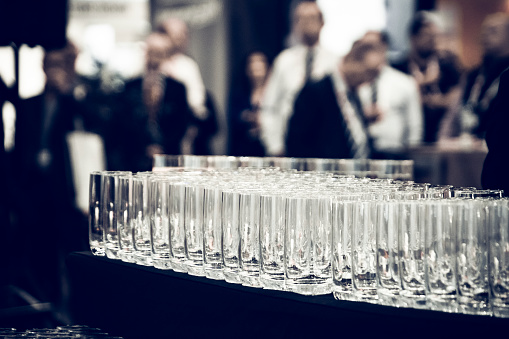 Rows of empty tall drinking glasses in a bar waiting to be filled with drinks for thirsty conference and convention participants seen out of focus in the background.  Horizontal, copy space, available light, high ISO, dramatic processing