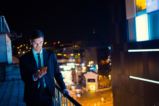 Young businessman at the rooftop of a skyscraper working late hours outside using his mobile phone