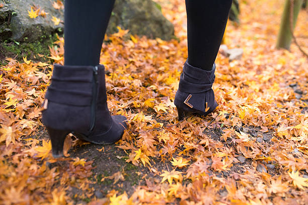 The legs of a woman in black pants and patent leather shoes stand on the grass with fallen maple leaves. Autumn theme.