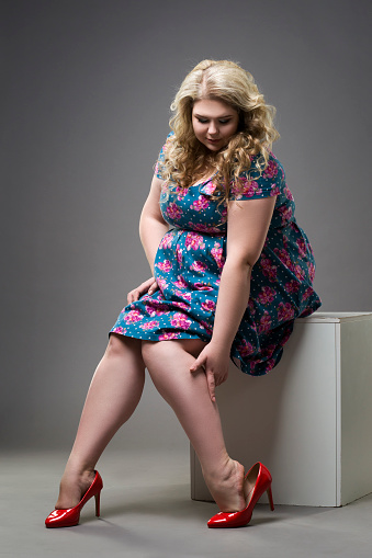Young beautiful blonde plus size model in red high heels shoes, xxl woman on gray studio background, legs fatigue