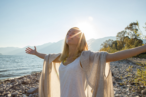 Portrait of young cheerful woman by the lake enjoying nature. Arms outstretched for positive emotion. 