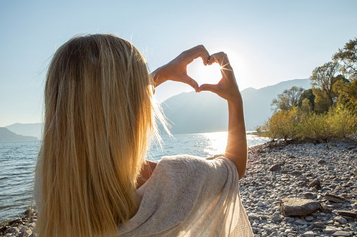 Young woman by the lake making a heart shape with her hands. Lake and mountain landscape, Swiss Alps on the background.