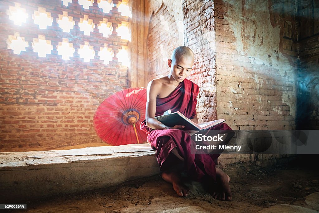 Burmese Novice Buddhist Monk in Temple Reading Bagan Myanmar Burmese buddhist Monk in his traditional clothing sitting inside temple reading buddhist book in the light of sunbeams shining through the ornamental temple window. Real People, Natural Interior Temple Light. Bagan, Mandalay Region, Myanmar, Asia. Monk - Religious Occupation Stock Photo