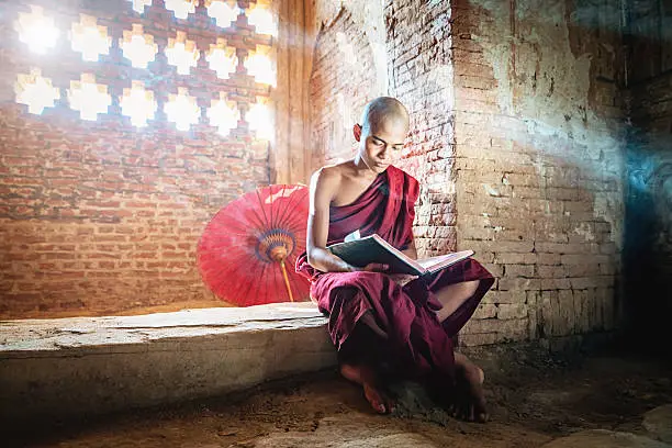 Burmese buddhist Monk in his traditional clothing sitting inside temple reading buddhist book in the light of sunbeams shining through the ornamental temple window. Real People, Natural Interior Temple Light. Bagan, Mandalay Region, Myanmar, Asia.