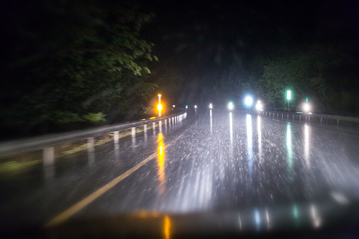Intense car driver personal perspective POV night shot of a severely motion blurred expressway highway driving lane during a drenching rain storm downpour looking through the windshield of a speeding car with its headlight high beams brightly illuminating an ominous string of warning road marker reflectors, the streaking guardrails along the edge of the highway, and the sloppy, slippery, dangerous, curving road ahead.