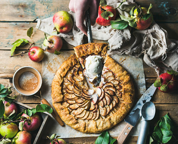 Man's hand holding piece of apple crostata pie Man's hand holding piece of apple crostata with cinnamon and ice-cream scoop over whole round pie served with fresh garden apples with leaves on rustic wooden background, top view, selective focus crostata stock pictures, royalty-free photos & images