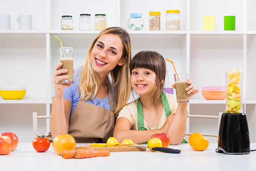 Happy mother and her daughter enjoy making and drinking smoothies together at their home.