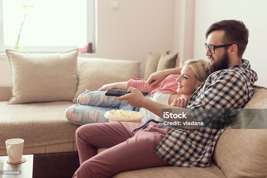 Couple watching a movie Young couple relaxing in their apartment, lying on the couch, watching a movie and eating popcorn. Lens flare effect on the window, focus on the guy Television Set Stock Photo