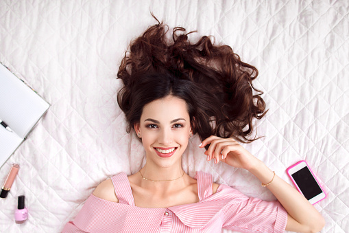 Beautiful woman lying on bed top view. Happy brunette in pink with healthy perfect hair smiling at camera. Beauty, joy, health care, fashion concept