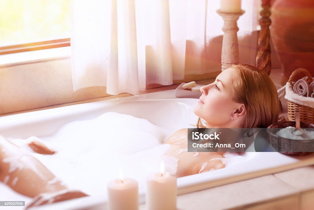 Enjoying day spa Beautiful woman lying down in the bath with foam and looking at the window, relaxation at home, hygiene and health care, happy and peaceful day spa Bathtub Stock Photo