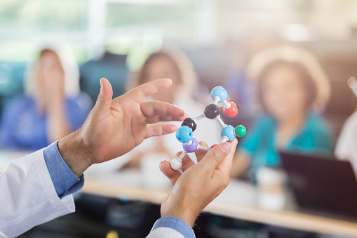 Male medical school professor uses a molecular structure model during lecture. He is showing the class a part of the model. Close-up is on the professor's hands holding the model. The class is blurred in the background.