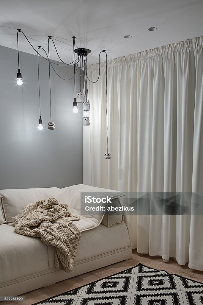 Room in modern style Interior in a modern style with gray wall, white ceiling and a parquet with a carpet on the floor. There is a beige sofa with a brown plaid, light curtains, hanging glowing lamps and decorative owls. Curtain Stock Photo