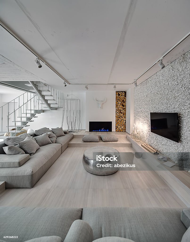 Interior in modern style Contemporary interior with white walls. There is white stair with a metal railing, sofas with pillows, sparkle pouf, burning candlesticks and fireplace, TV, walls decorated with firewood and pebbles. Apartment Stock Photo