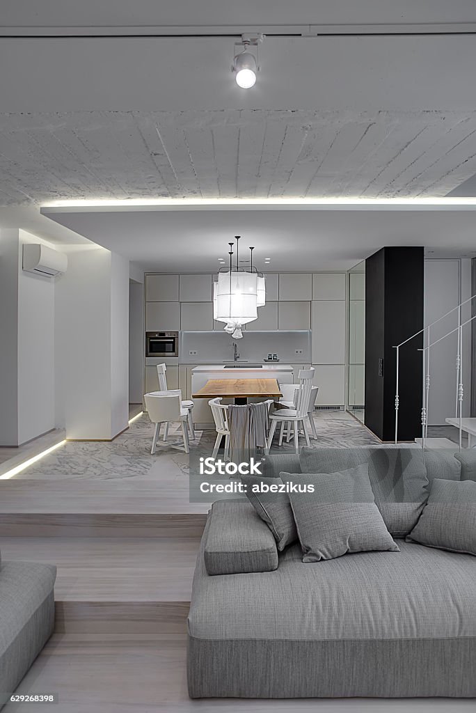 Interior in modern style Modern interior with white walls and a stair with metal railing. There is a gray sofa with pillows, kitchen zone with wooden table and chairs, glowing lamps with white lampshades. Vertical. Apartment Stock Photo