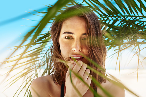 Shot of an attractive young woman standing under the shade of a palm tree at the beach