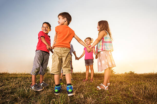 A multi-ethnic group of elementary age children standing together in a row, holding hands with their arms raised in the air, smiling and looking at the camera. Standing atop a grassy hill with the clear blue sky in the background.