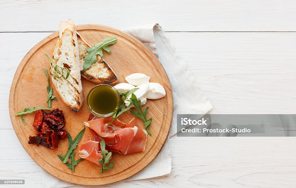 Catering platter antipasto with prosciutto and mozzarella Catering platter antipasto with ham prosciutto, bruschetta bread toasts and mozzarella cheese on round wooden board on white wood table top view with copy space. Served starter meals, restaurant food Antipasto Stock Photo