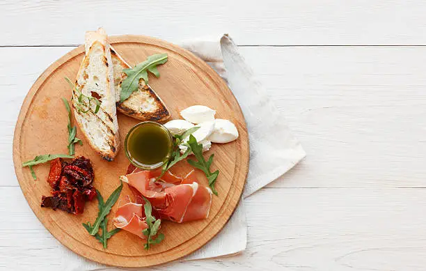 Catering platter antipasto with ham prosciutto, bruschetta bread toasts and mozzarella cheese on round wooden board on white wood table top view with copy space. Served starter meals, restaurant food