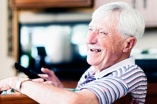 Laughing 90-year-old old man is happily relaxing by watching television, smiling over his shoulder at camera as he holds the remote control,  changing channnels as he pleases. Copy space on the defocused TV screen.