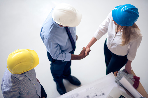 Architect and businesswoman shaking hands on construction site. They are wearing business clothes and protective helmets. Another businessman stands next to them. Shot from above.