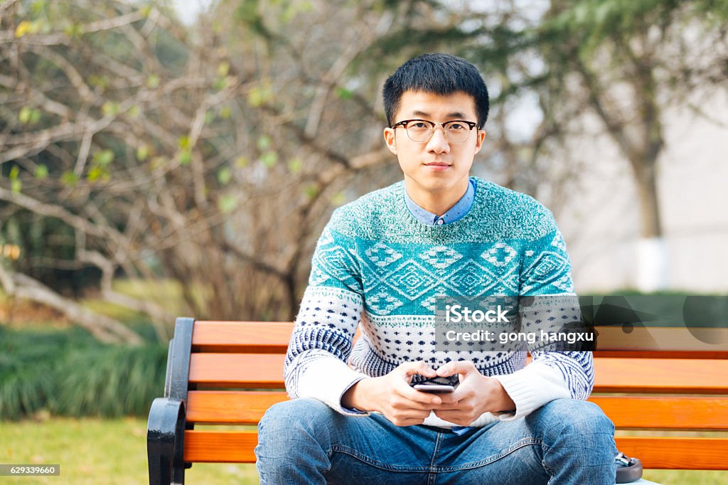 young asian man holds mobile phone young asian man holding mobile phone sits on long bench in park Adult Stock Photo