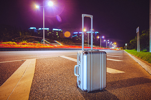 Night shot of aluminium suitcase beside the highway. Visible city lights in the background. Horizontal shot, lots of copyspace.